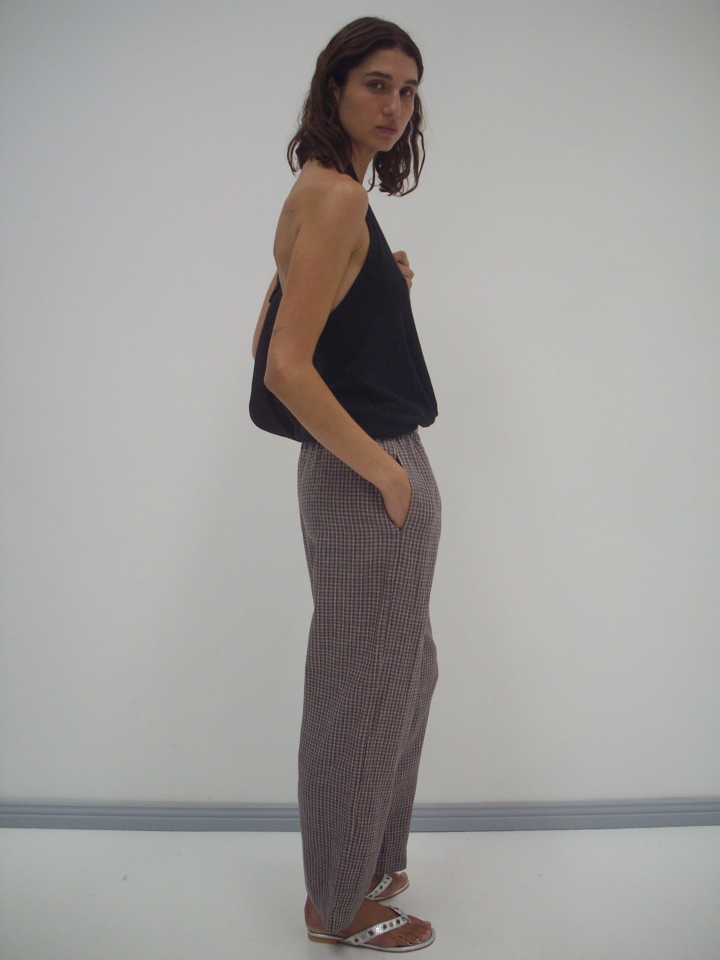 A person with shoulder-length brown hair stands sideways by a white wall, wearing a black Jersey Top - Black from Deiji Studios, plaid wide-leg pants, and brown sandals, with one hand in their pocket.