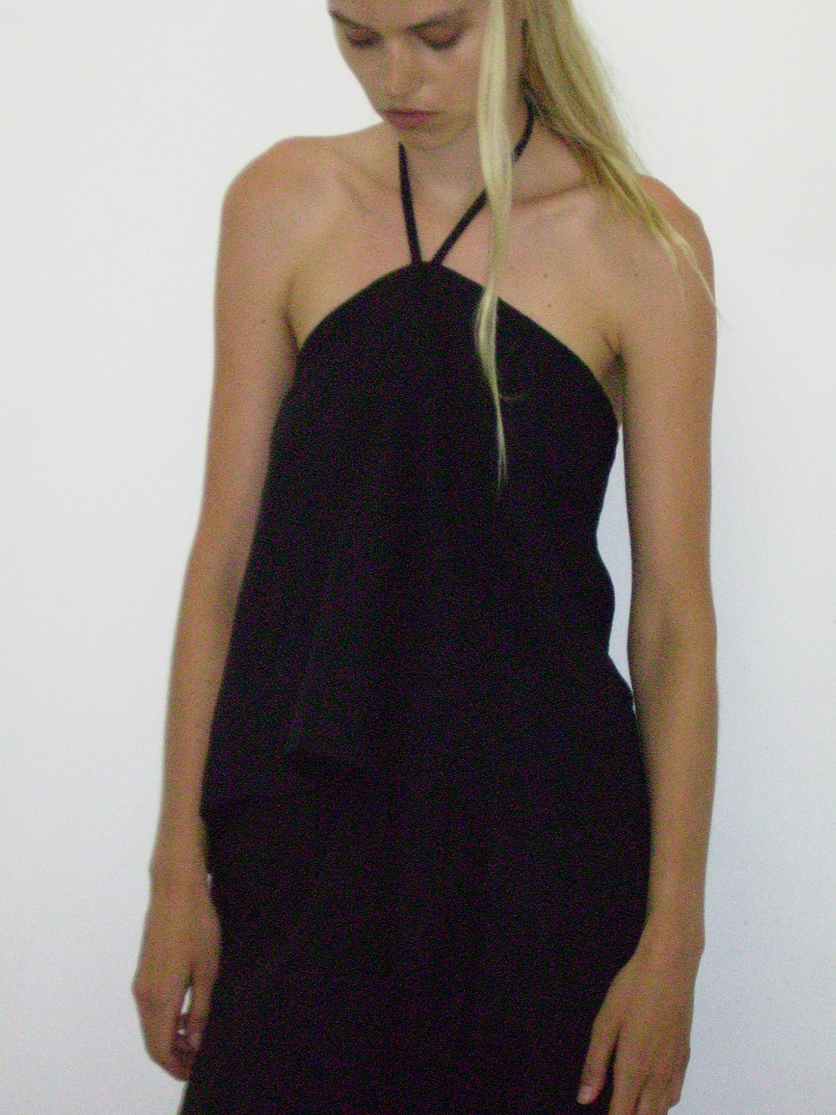 A woman with long blonde hair wears the Linen Wrap Back Top - Black from Deiji Studios, made of soft French linen. She stands against a plain white background, looking down with arms relaxed at her sides.