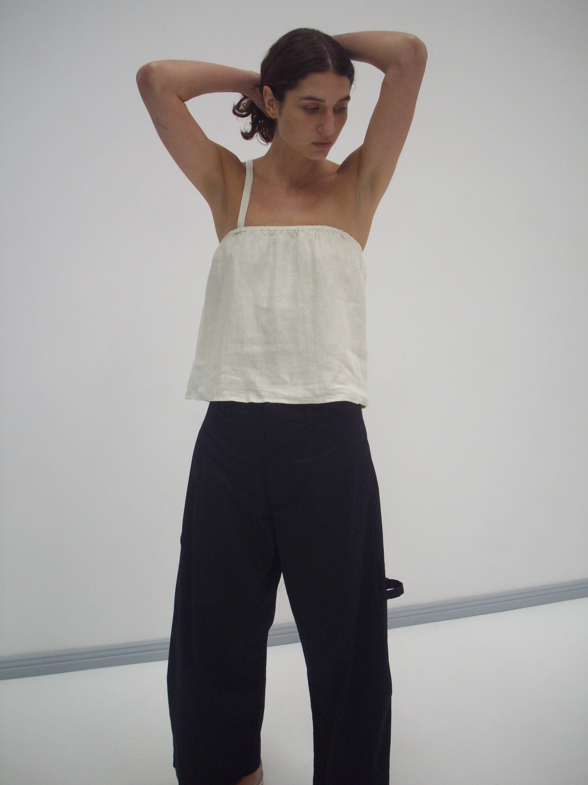 A person wears a Button Strap Top in Pastel Pistachio and loose black pants, standing against a plain white background with arms raised behind their head, looking down thoughtfully.