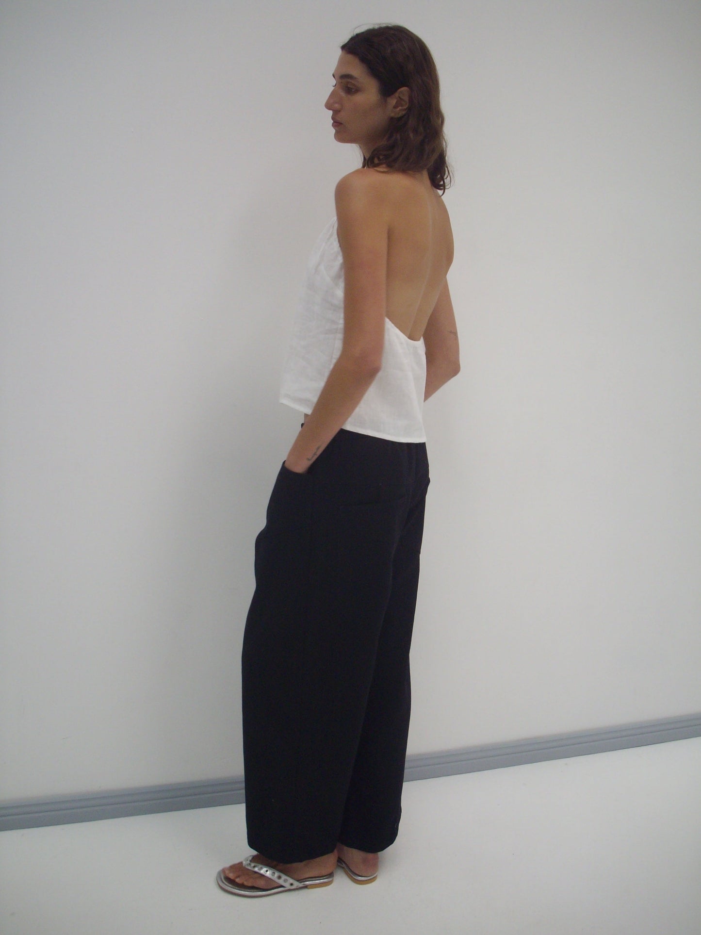 A person stands sideways against a white wall, wearing the Square Pocket Pant - Black with a white backless halter top and flat sandals, hands in pockets, looking slightly over their shoulder.