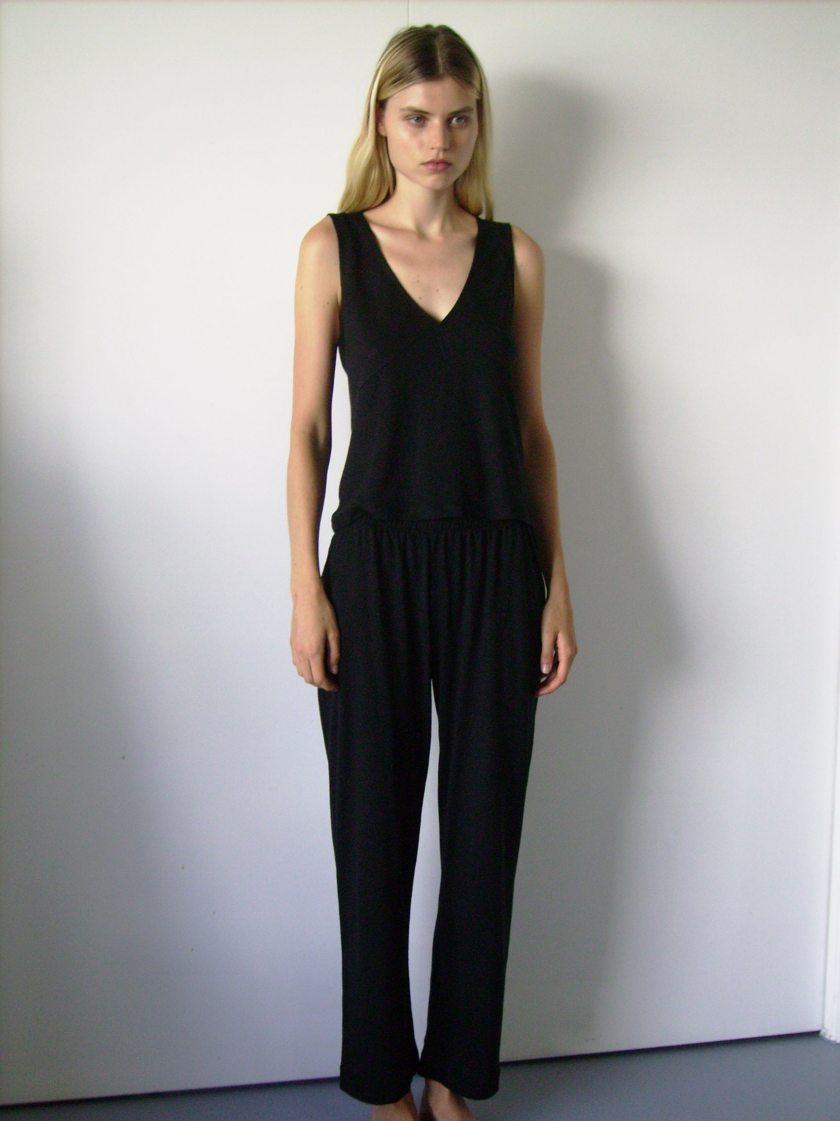 A woman with long blonde hair stands barefoot on a blue floor against a white wall, wearing Deiji Studios Soft Pant and a sleeveless black top. Her arms are rested beside her and has a neutral expression.