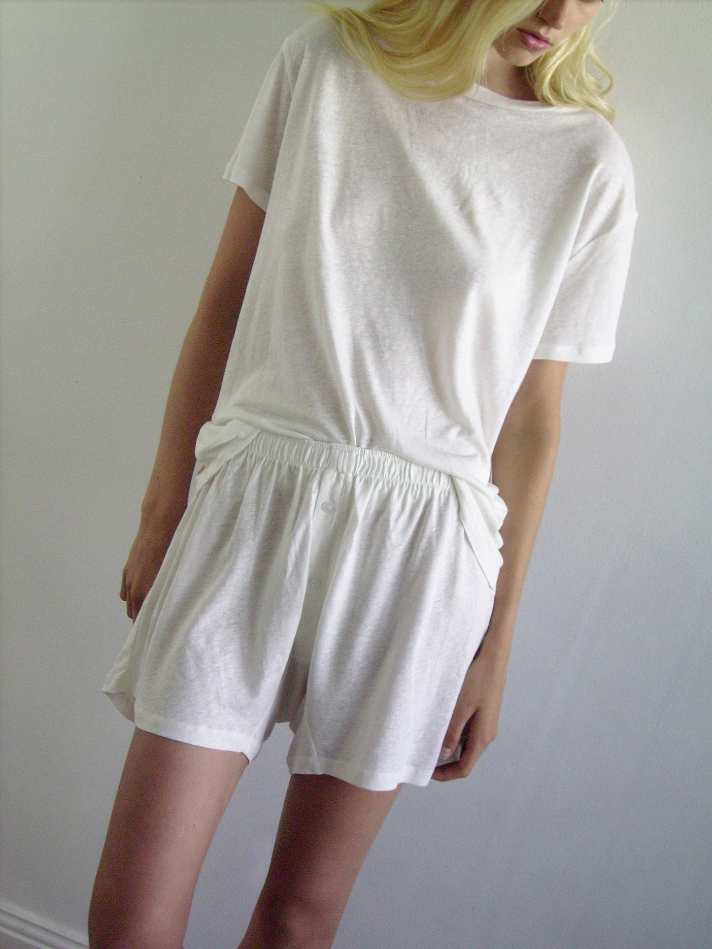 A person with long blonde hair wears a loose white t-shirt and Soft Short, standing against a plain white wall. Their head is slightly tilted, and their face is partially out of frame.