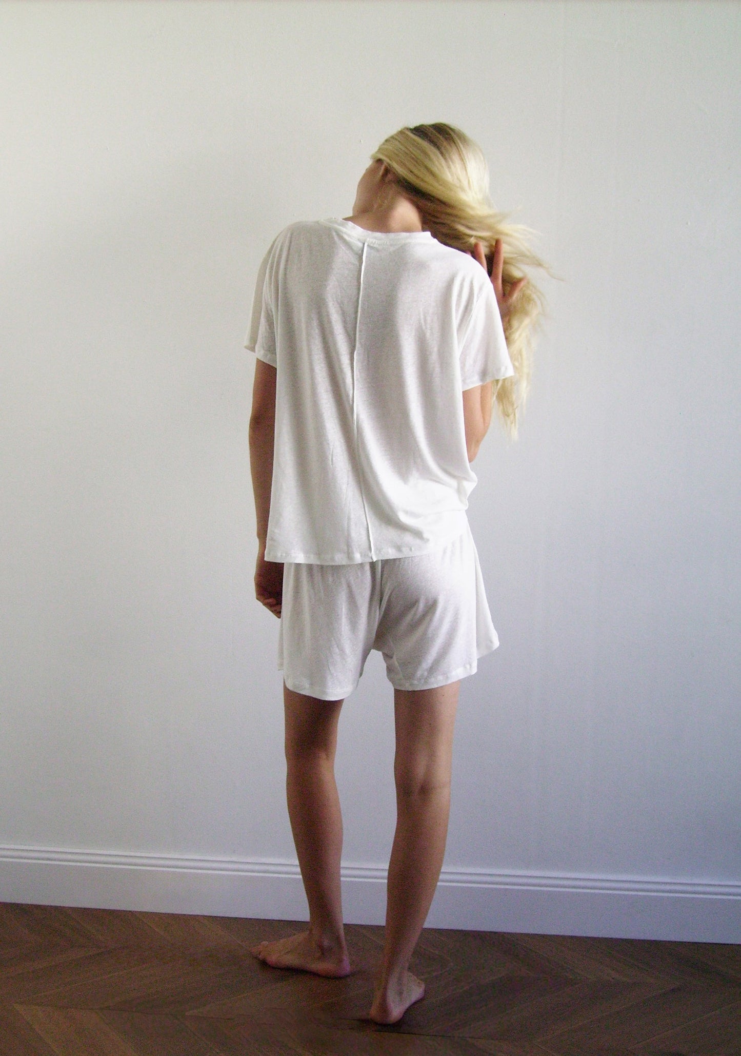 A person with long blonde hair, wearing 0s soft t shirt - ecru and matching shorts, stands barefoot on a wooden floor facing a plain white wall, head turned and hand touching their hair.