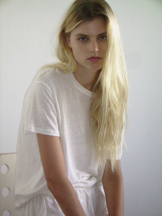 A person with long blonde hair and light skin sits on a white chair wearing the Deiji Studios soft t shirt in ecru and loose white pants, looking at the camera with a neutral expression against a plain background.