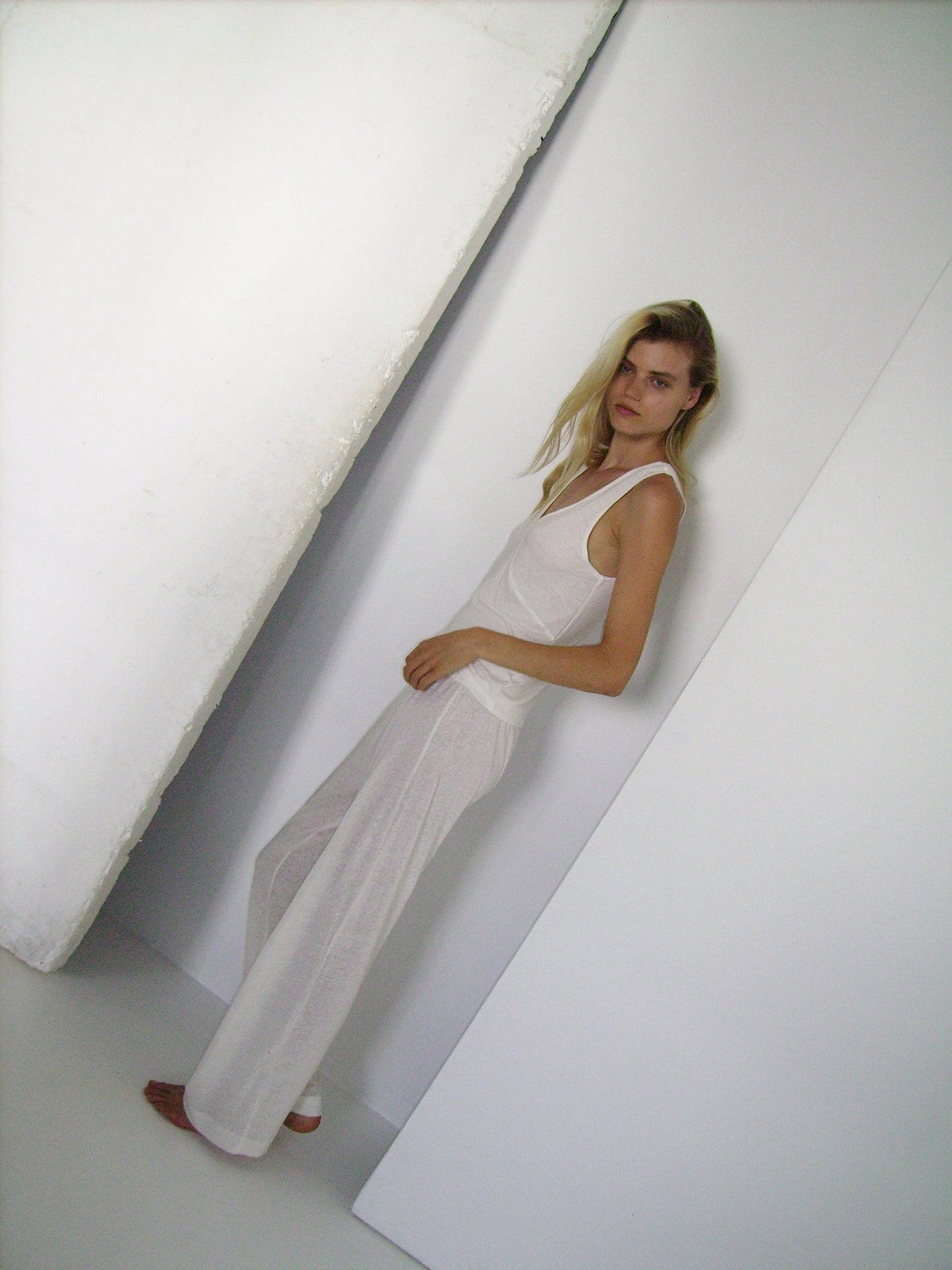 A woman with long blonde leans against a white wall, wearing a loose-fitting, sleeveless white top and 0’s soft pant - ecru loungewear. She is looking directly at the camera with a neutral expression