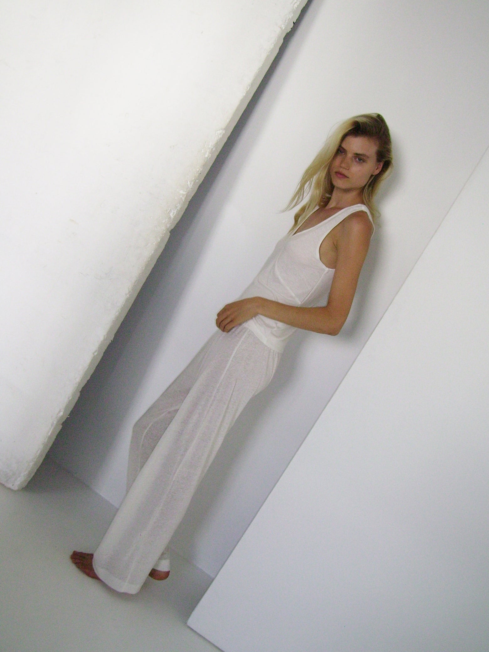 A woman stands barefoot against a white wall, wearing the Deiji Studios soft tank - ecru and loose white loungewear pants, looking neutrally at the camera in a minimalistic, bright space.
