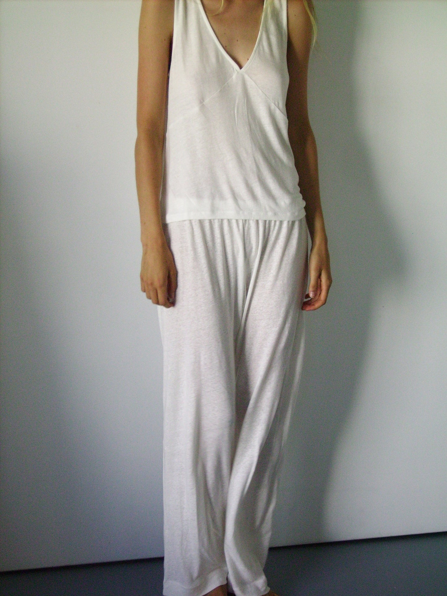 A woman stands upright against a white wall, wearing the Deiji Studios soft tank - ecru and loose white loungewear pants, her head is cut out of the frame.