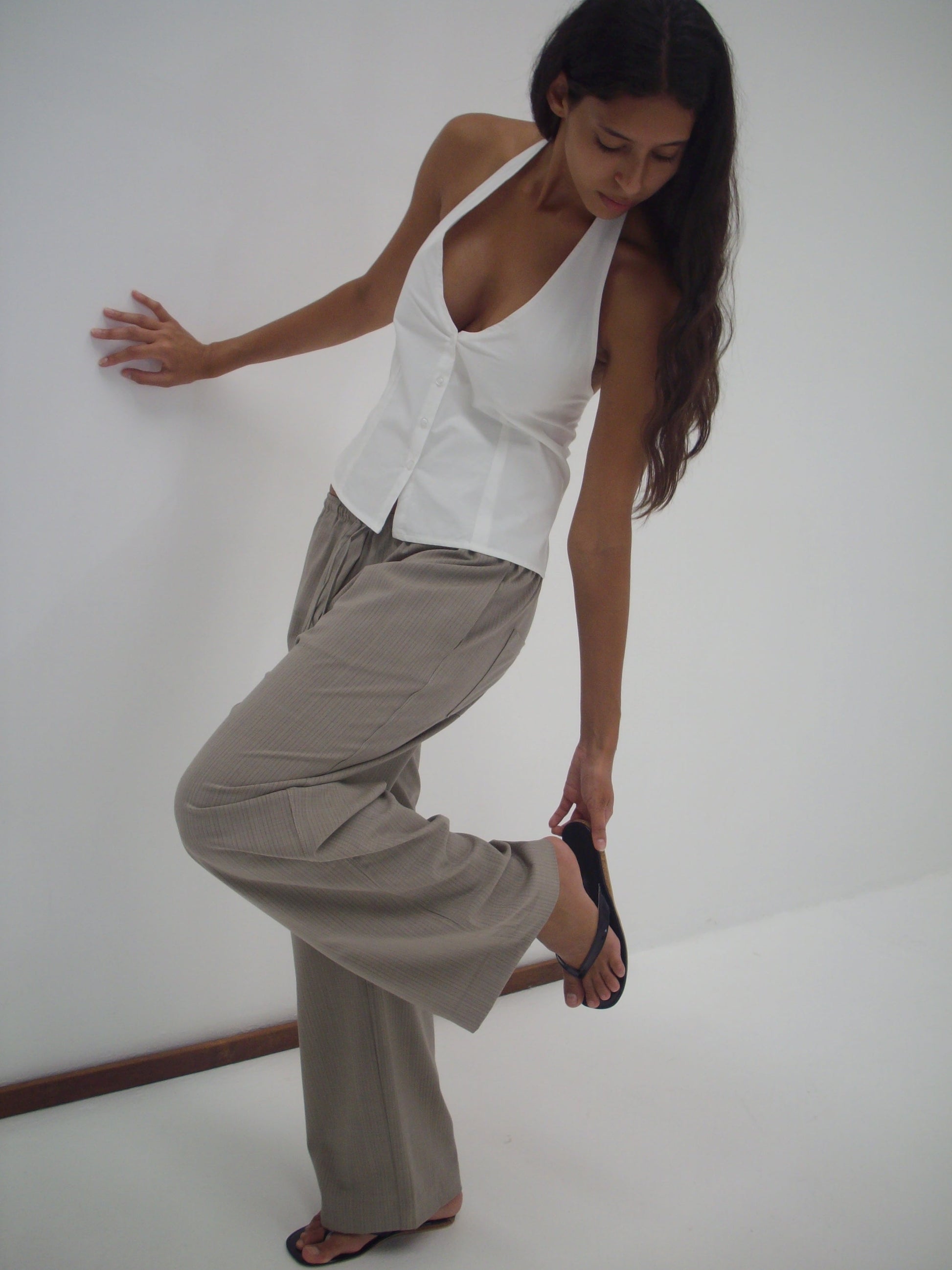 A woman wearing the Low Pocket Pure Wool Pant - Dove Stripe, a white halter top, and black sandals stands on one foot against a white wall and floor, looking down as she adjusts her sandal.