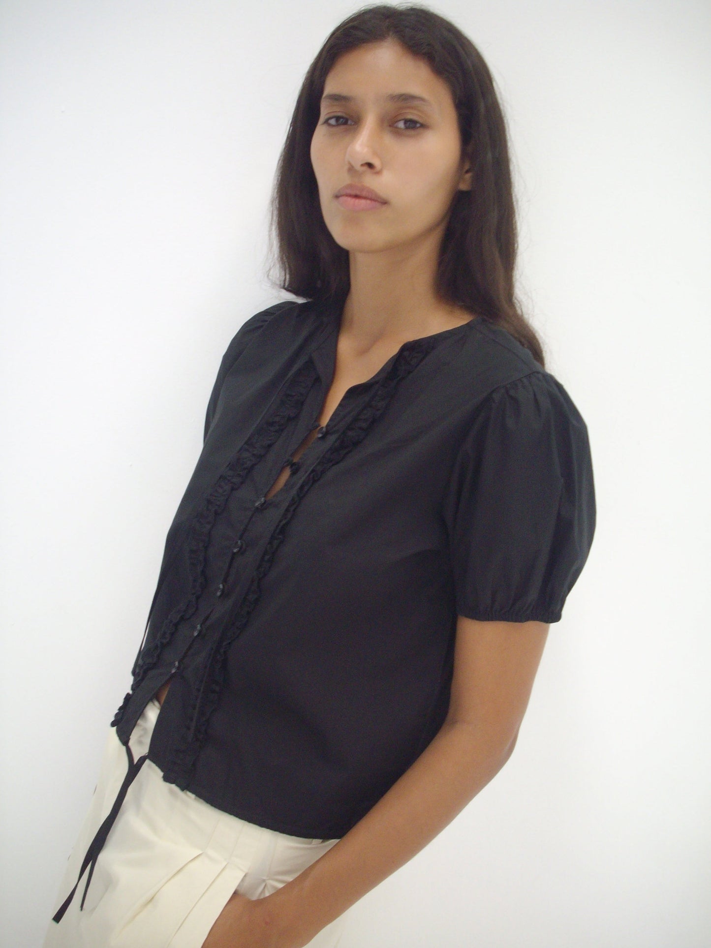 A woman with long dark hair wears the Double Frill Top, a black short-sleeved blouse, paired with a white skirt. She stands against a plain white background and looks at the camera with a neutral expression.