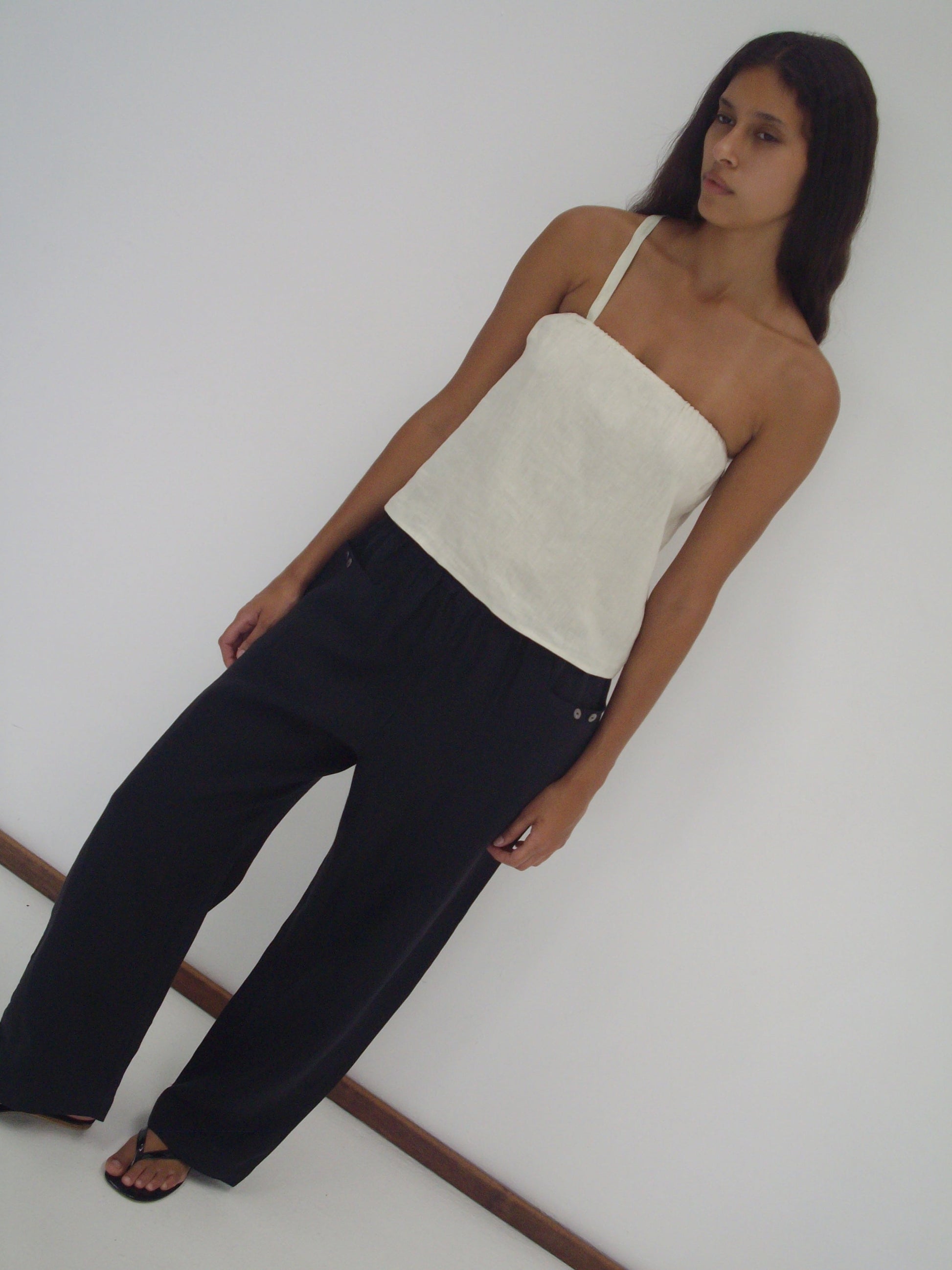 A woman with long dark hair stands against a white wall, wearing the Button Strap Top - Pastel Pistachio by Deiji Studios in organic French linen, paired with loose black pants and black sandals, her expression remains neutral.