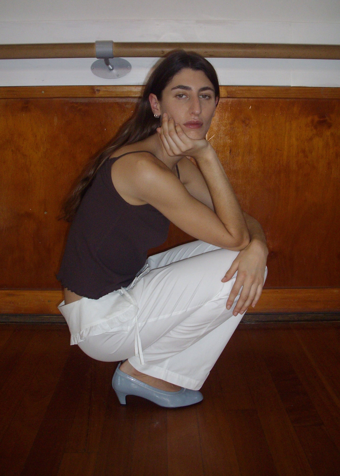 A person with long brown hair squats indoors by a wooden wall, resting their chin on one hand. They wear a dark sleeveless top, Flounce Belt Pant - White by Deiji Studios, and light blue heels, gazing calmly at the camera.