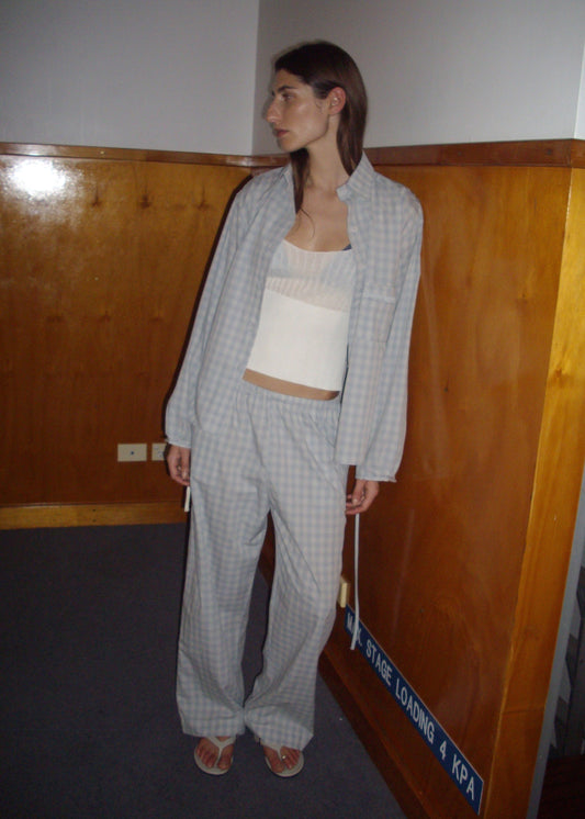 A woman stands indoors in Deiji Studios’ Side Tie Pant – Blue Check and a loose light gray plaid jacket over a white top and sandals, facing sideways toward a wood-paneled wall and a blue "STAGE LOADING 4 KPA" sign.
