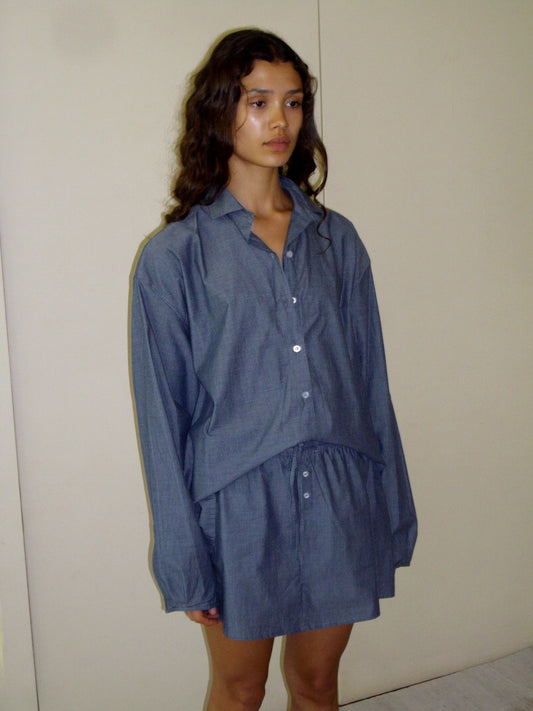 A woman with long wavy hair stands against a plain wall, wearing the day shirt in blue with a matching skirt. She looks slightly to the side, her expression neutral.