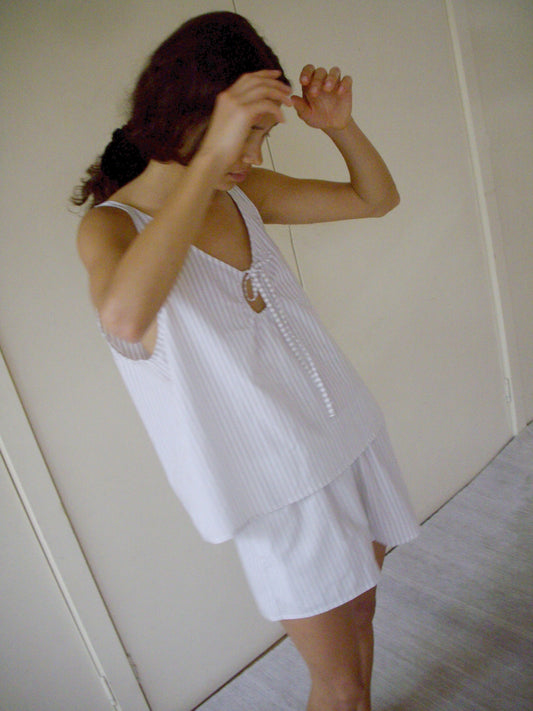 Someone stands indoors with arms raised, partially covering their face, wearing the U Shape Cotton Set—a loose, sleeveless top and matching shorts with light stripes. A light-colored wall and door are visible in the background.