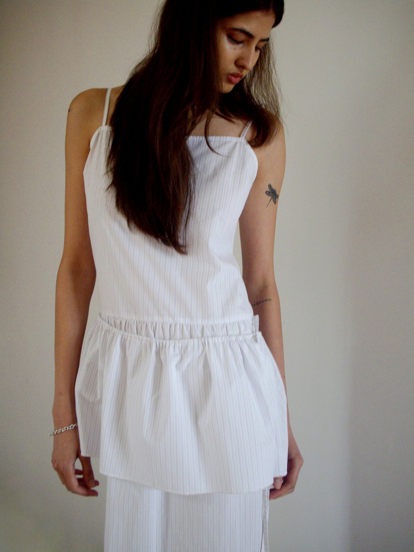 A person with long brown hair wears the 0 Peplum Dress - Pacific Stripe, a sleeveless style with a gathered waist. They look down with one hand at their side, revealing a dragonfly tattoo on their upper arm against a light plain background.