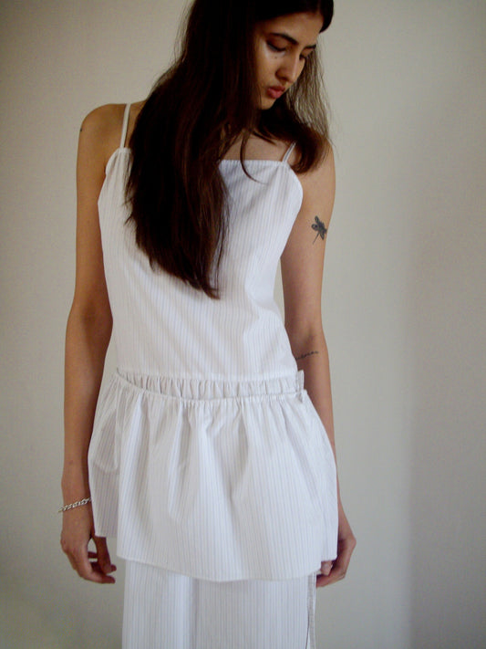 A person with long brown hair wears the 0 Peplum Dress - Pacific Stripe, a sleeveless style with a gathered waist. They look down with one hand at their side, revealing a dragonfly tattoo on their upper arm against a light plain background.