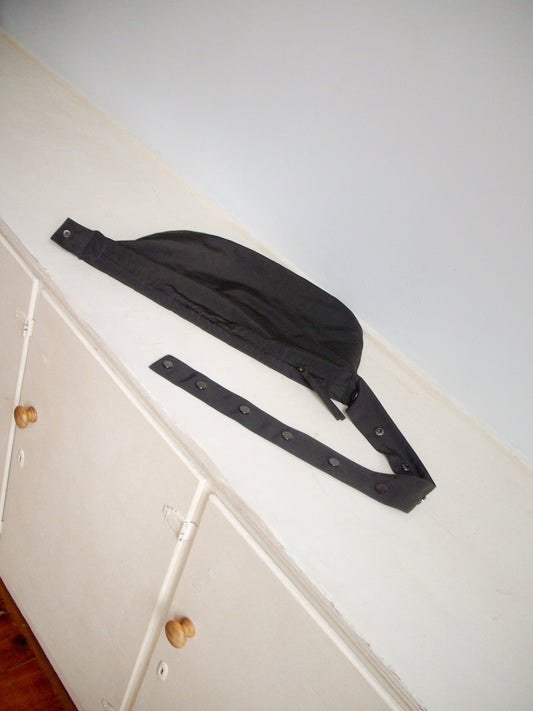 A Press Stud Saddle Bag in dark chocolate recycled polyester sits atop a white cabinet with wooden knobs, set against a plain light gray wall.