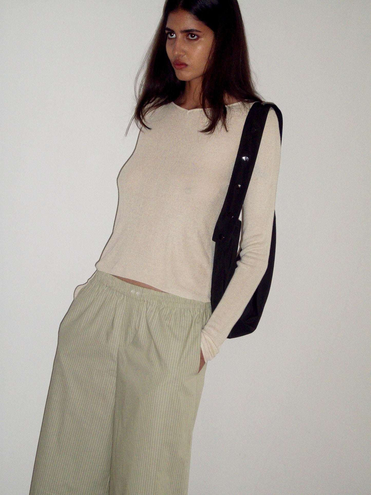 A woman with long brown hair stands against a white wall, wearing the 0 Knitted Top - Fawn and loose light green striped pants. She has a black bag over her shoulder, hands in pockets, and looks to the side.