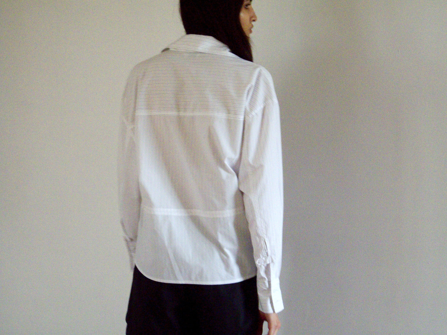 A person with long dark hair, seen from behind, wears the 0 Room Shirt - Pacific Stripe and black shorts while standing against a plain light-colored wall.