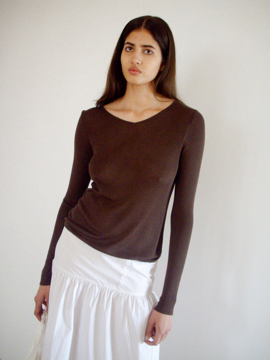 A woman with long dark hair wears the 0 Knitted Top - Dark Brown, a fitted, long-sleeved ecoviscose knit with sculptural seam details, styled with a white skirt as she stands against a plain light wall and calmly faces the camera.
