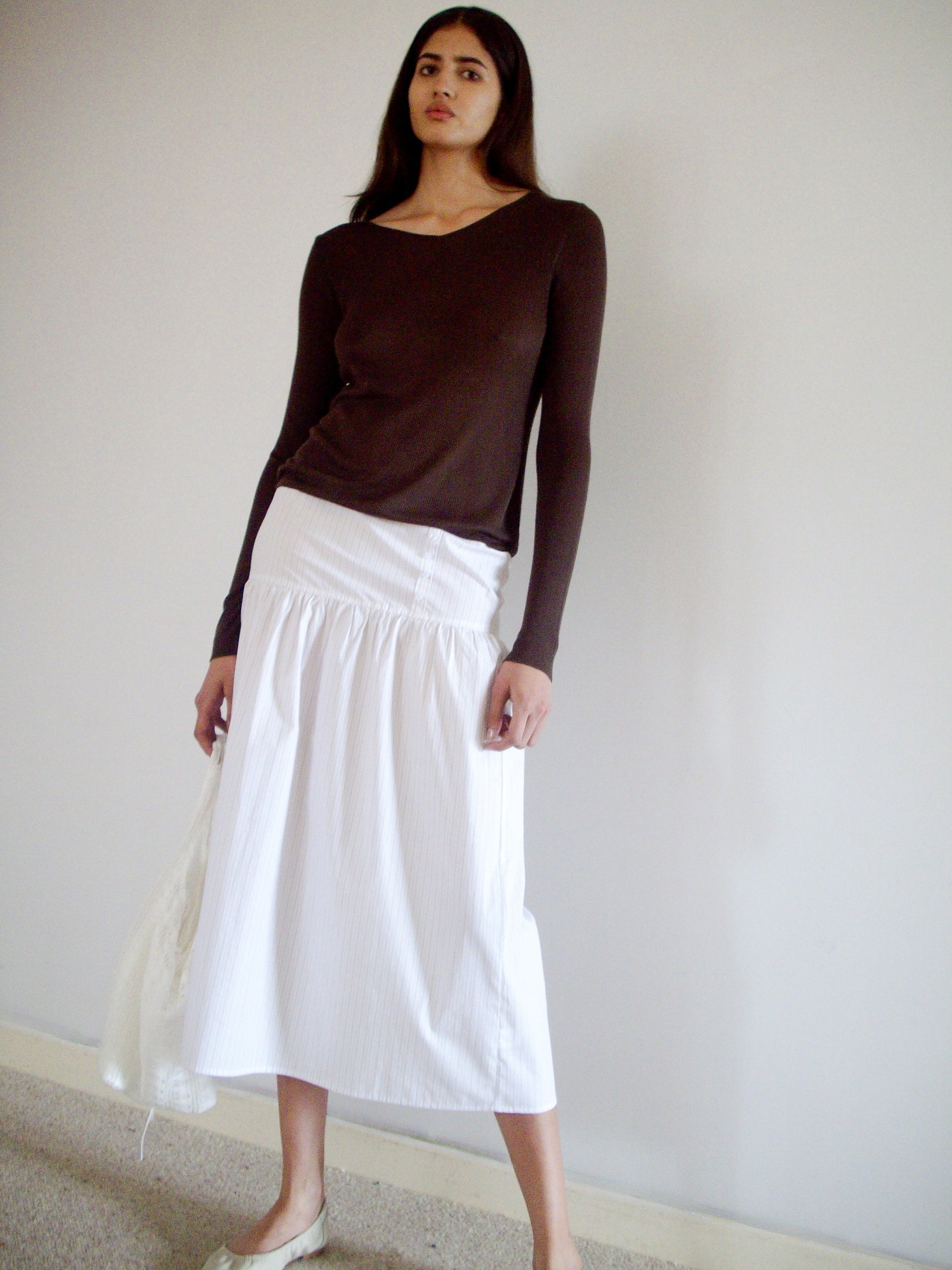 A woman with long dark hair stands indoors against a plain wall, wearing the 0 Knitted Top - Dark Brown, a long white skirt, and light-colored flats. She has a neutral expression.