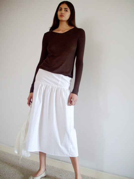 A woman with long dark hair stands indoors against a plain wall, wearing the 0 Knitted Top - Dark Brown, a long white skirt, and light-colored flats. She has a neutral expression.