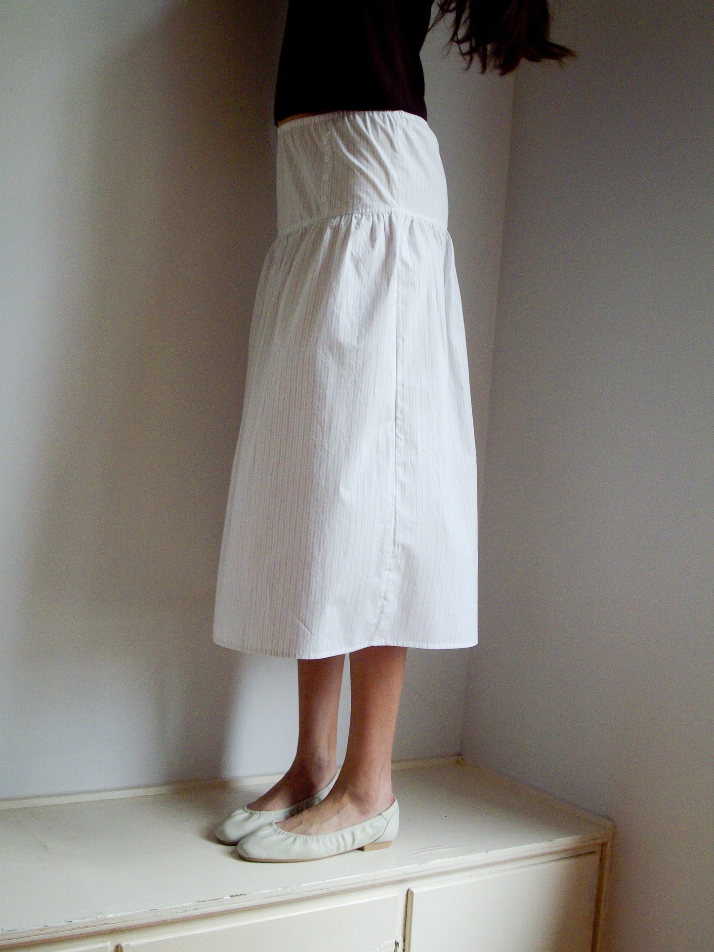 A person stands on a cream-colored cabinet, wearing the Button Skirt - Pacific Stripe by 0. The long skirt features an elasticated waistline; the upper body is mostly out of frame. The setting is indoors with plain walls and white shoes are worn.