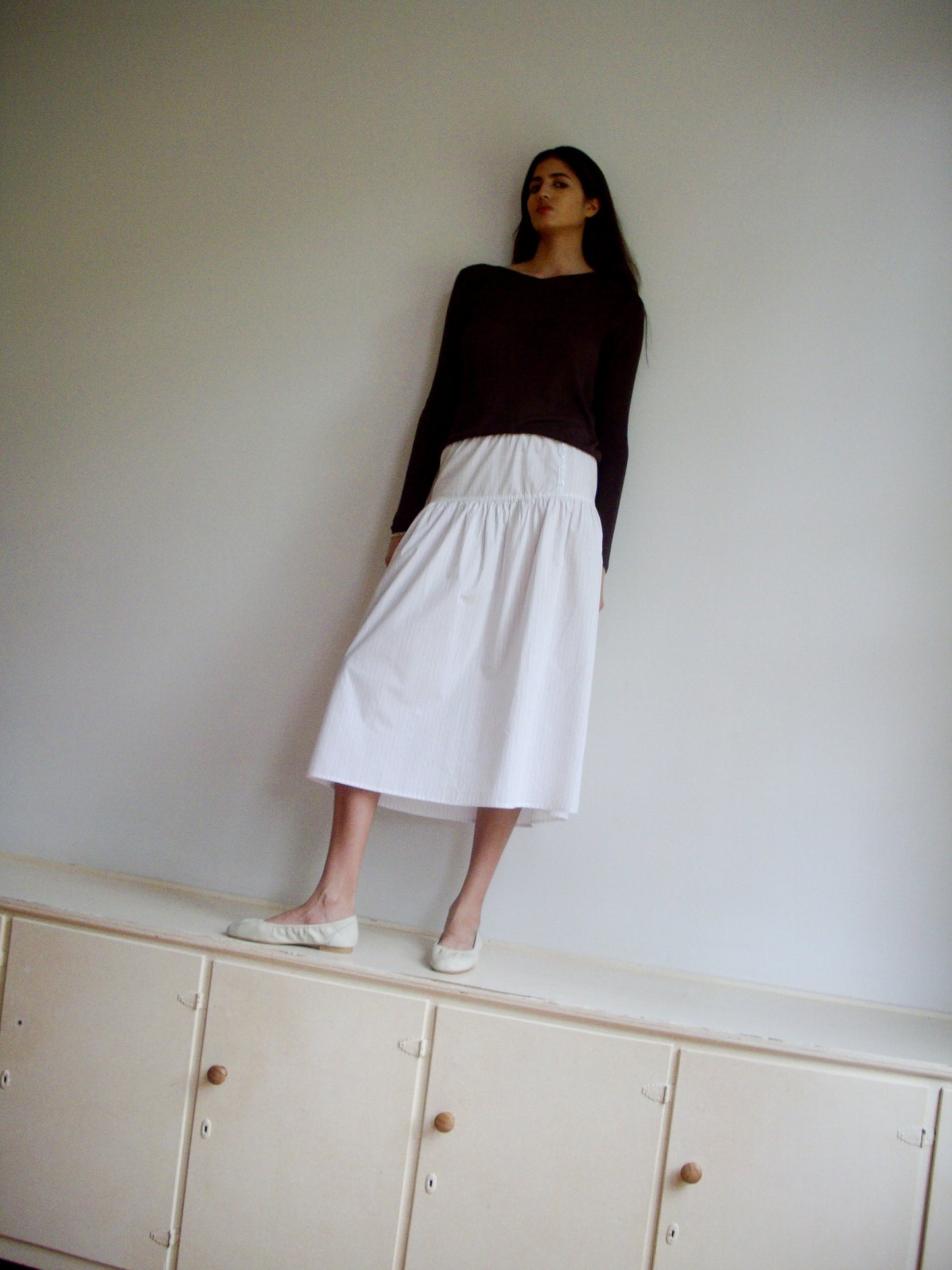 A woman in a black long-sleeve top and the Button Skirt - Pacific Stripe by 0, featuring an elasticated waistline, stands on a light wooden cabinet against a plain white wall, gazing slightly to the side.