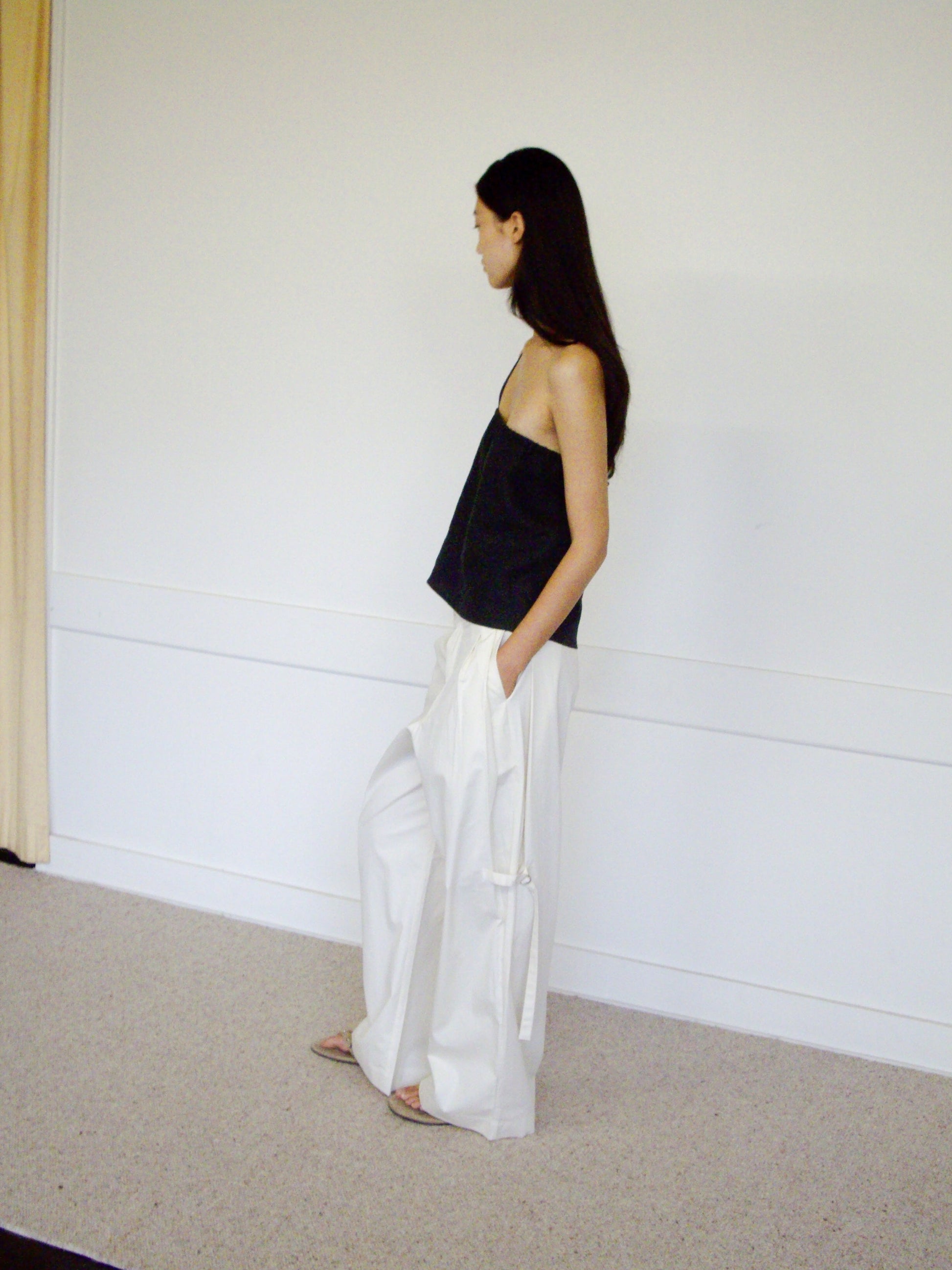 A woman with long dark hair stands sideways in a minimalist room, wearing the 0 Tab Pant - Off White, a loose black camisole with adjustable metal straps, and sandals.