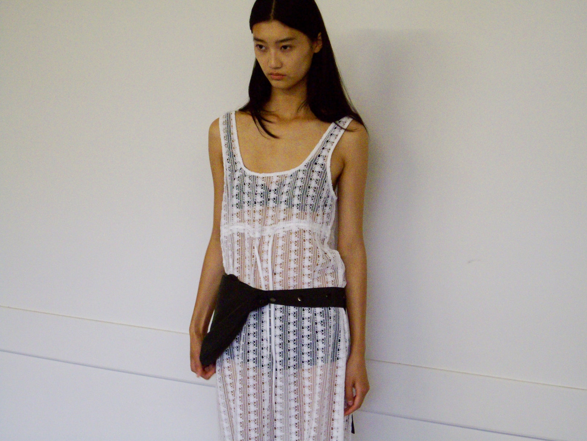 A woman with long dark hair stands against a plain wall, wearing the Thread Dress - Off White by 0 over a black undergarment and a black waist bag. She has a neutral expression, arms by her sides.