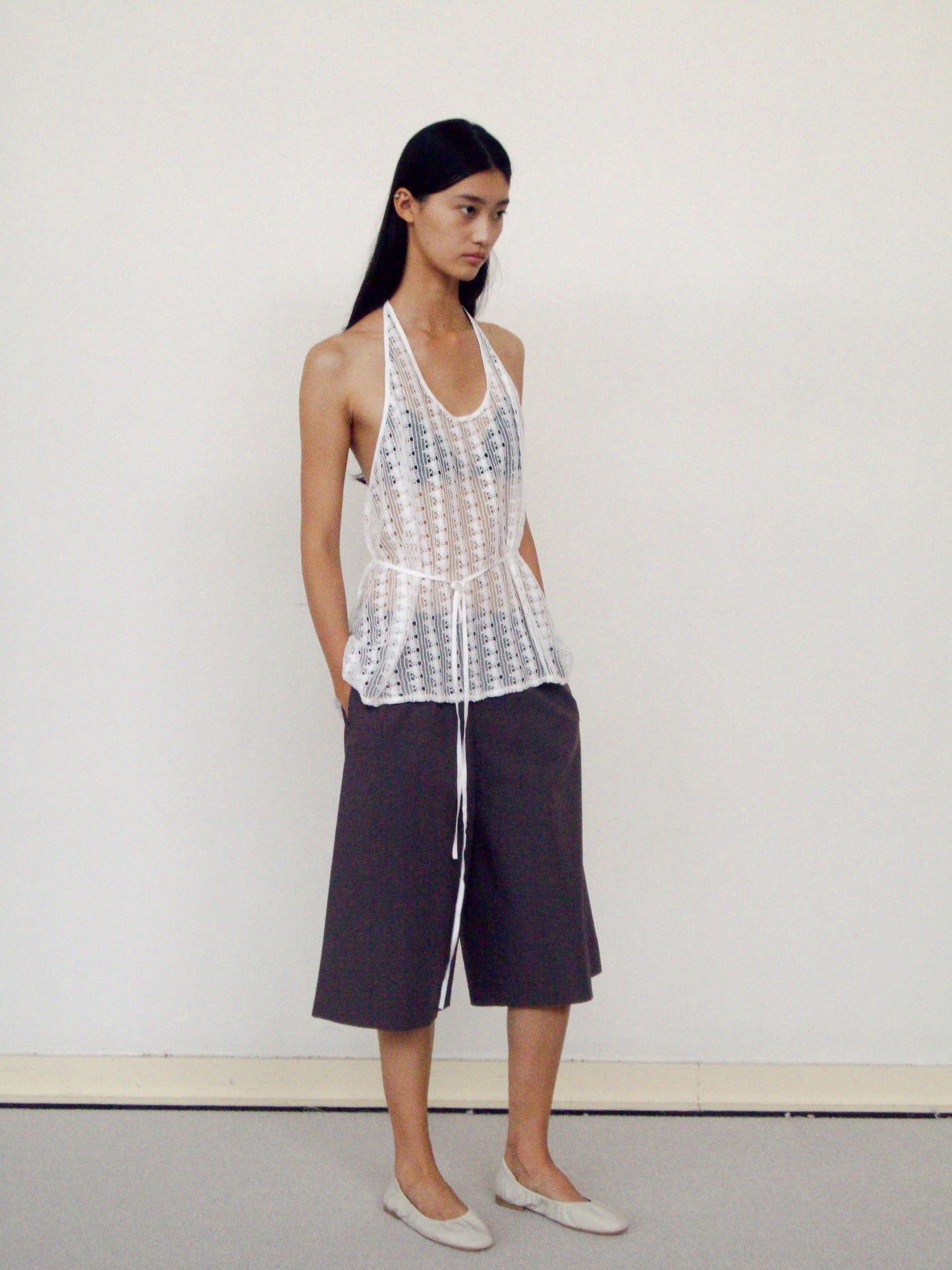 A woman with long straight hair stands against a plain wall, wearing the 0 Thread Wrap Top in Off White, loose dark gray culotte shorts, and light-colored flats, with her hands in her pockets.