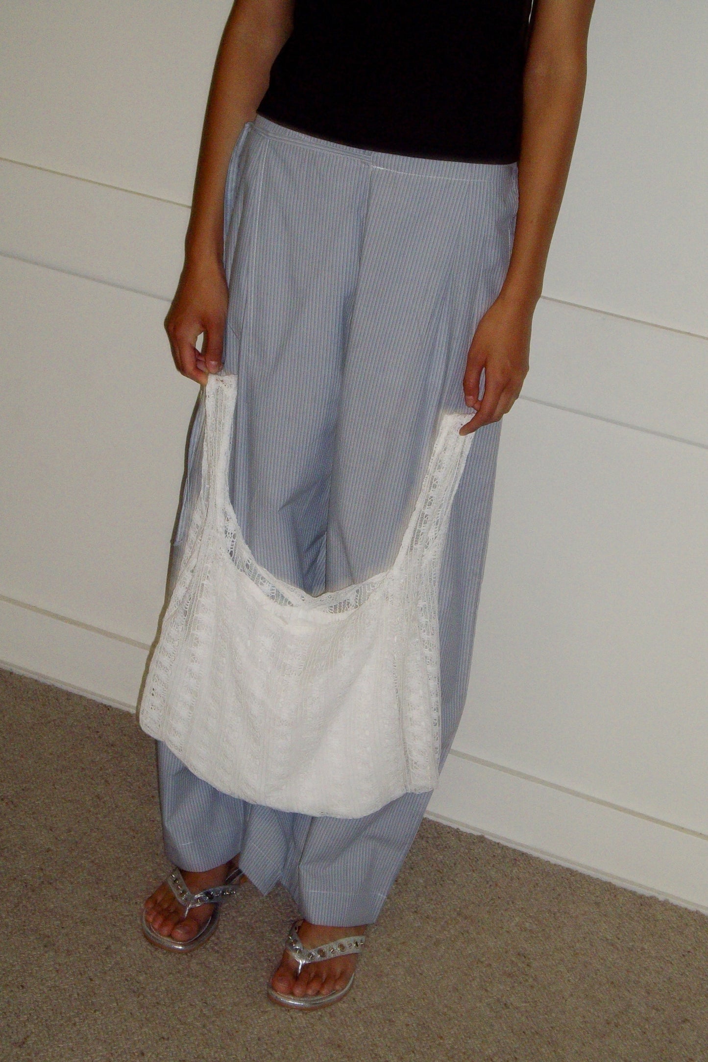 A person stands indoors wearing loose striped pants, a black top, and sandals, holding the Thread Dust Bag - Off White organic cotton tote from Deiji Studios in front of them. Their head is not visible in the image.