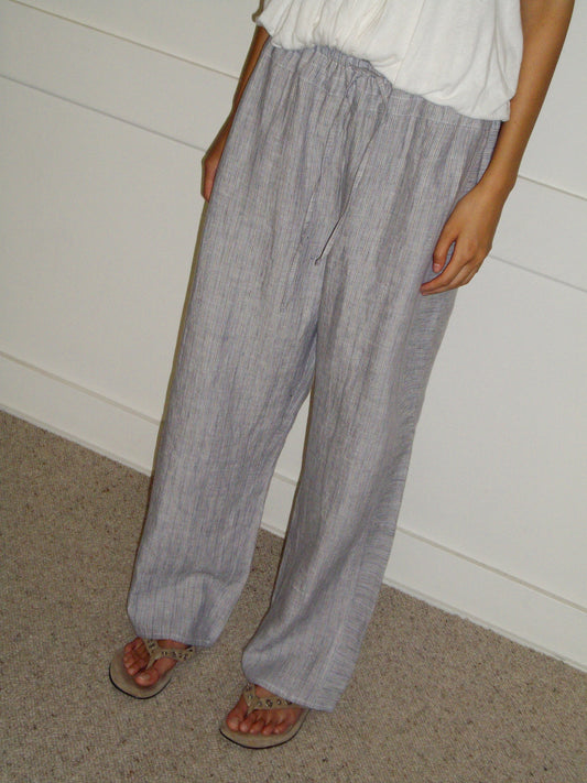 A person stands indoors wearing Deiji Studios’ Double Waist Linen Pant in Haze Blue, paired with a white top and sandals. Their hands rest by their sides, and the photo is cropped at the shoulders.