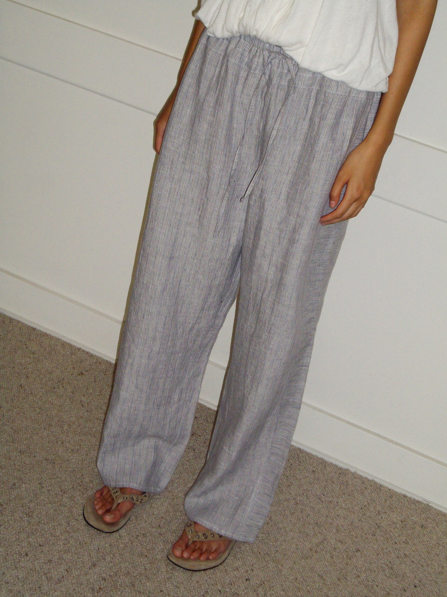 A person wearing 0s Double Waist Linen Pant in Haze Blue, paired with a white top and sandals, stands against a white wall and beige carpet. Their head is not shown in the image.