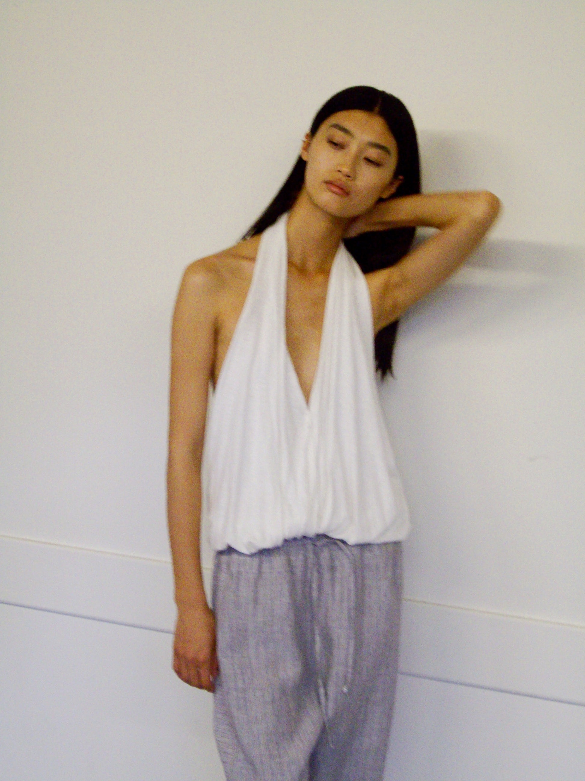A person with long dark hair wears the Jersey Top - White by 0, paired with light gray pants, standing against a plain wall with one hand resting on the back of their neck.