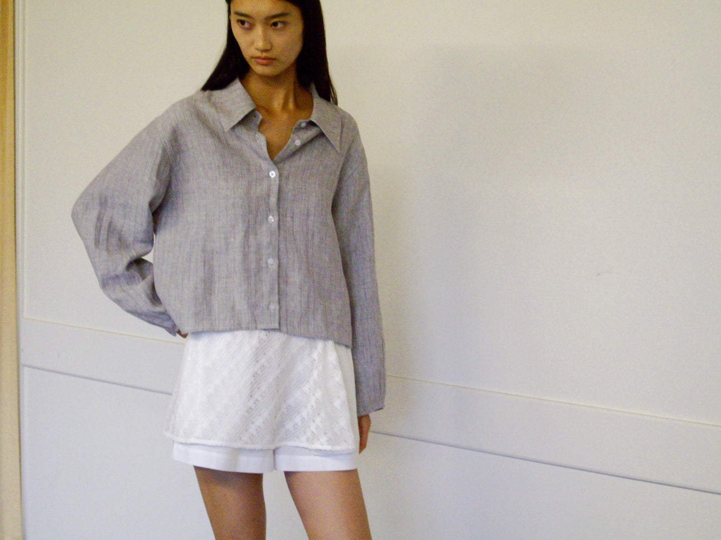 A woman with long dark hair and a neutral expression stands against a white wall, wearing the 0 Collar Point Shirt in Haze Blue over a white textured skirt, with one hand resting on her hip.