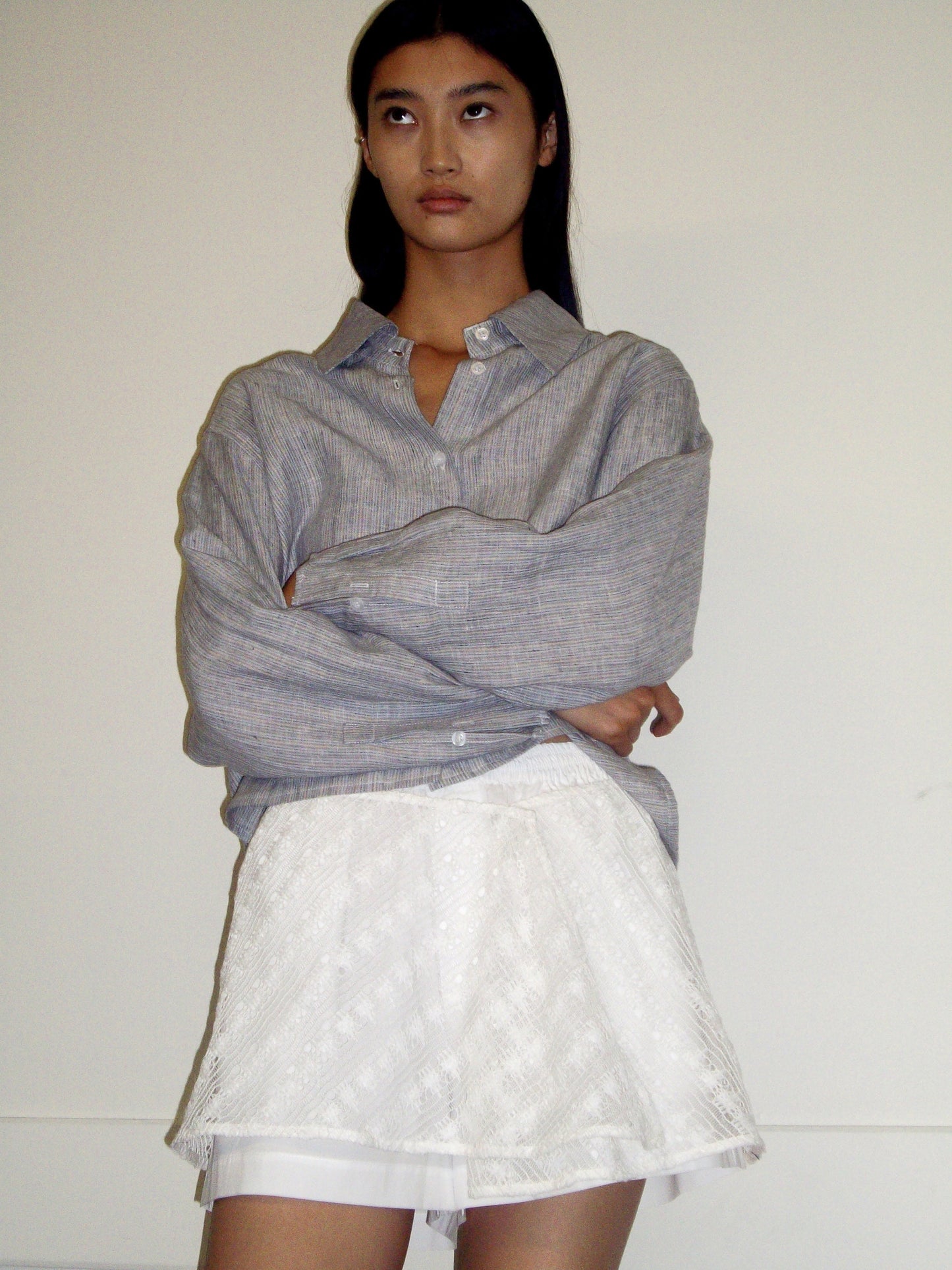 A person with long dark hair stands against a plain wall, wearing a loose gray button-up shirt and the Thread Button Belt - Off White as a short textured skirt. Their arms are crossed and their expression is neutral.