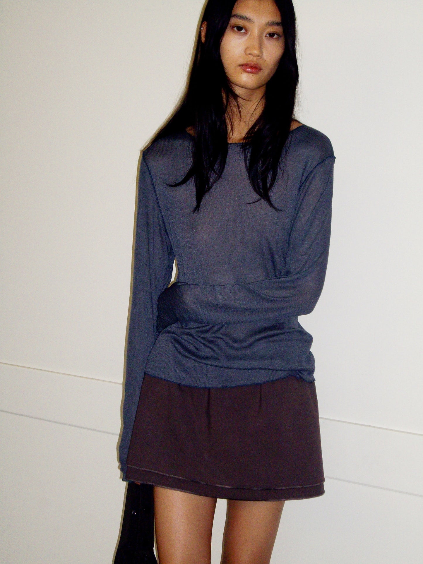 A person with long dark hair stands against a white wall, wearing a loose, long-sleeved gray organic cotton shirt and the Layered Skirt - Chocolate by 0, looking at the camera with a neutral expression.