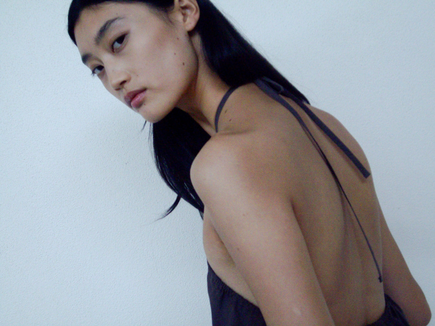 A person with long black hair models the Napkin Top - Chocolate by 0, a backless dark wrap halter top, looking over their shoulder toward the camera against a plain light background.