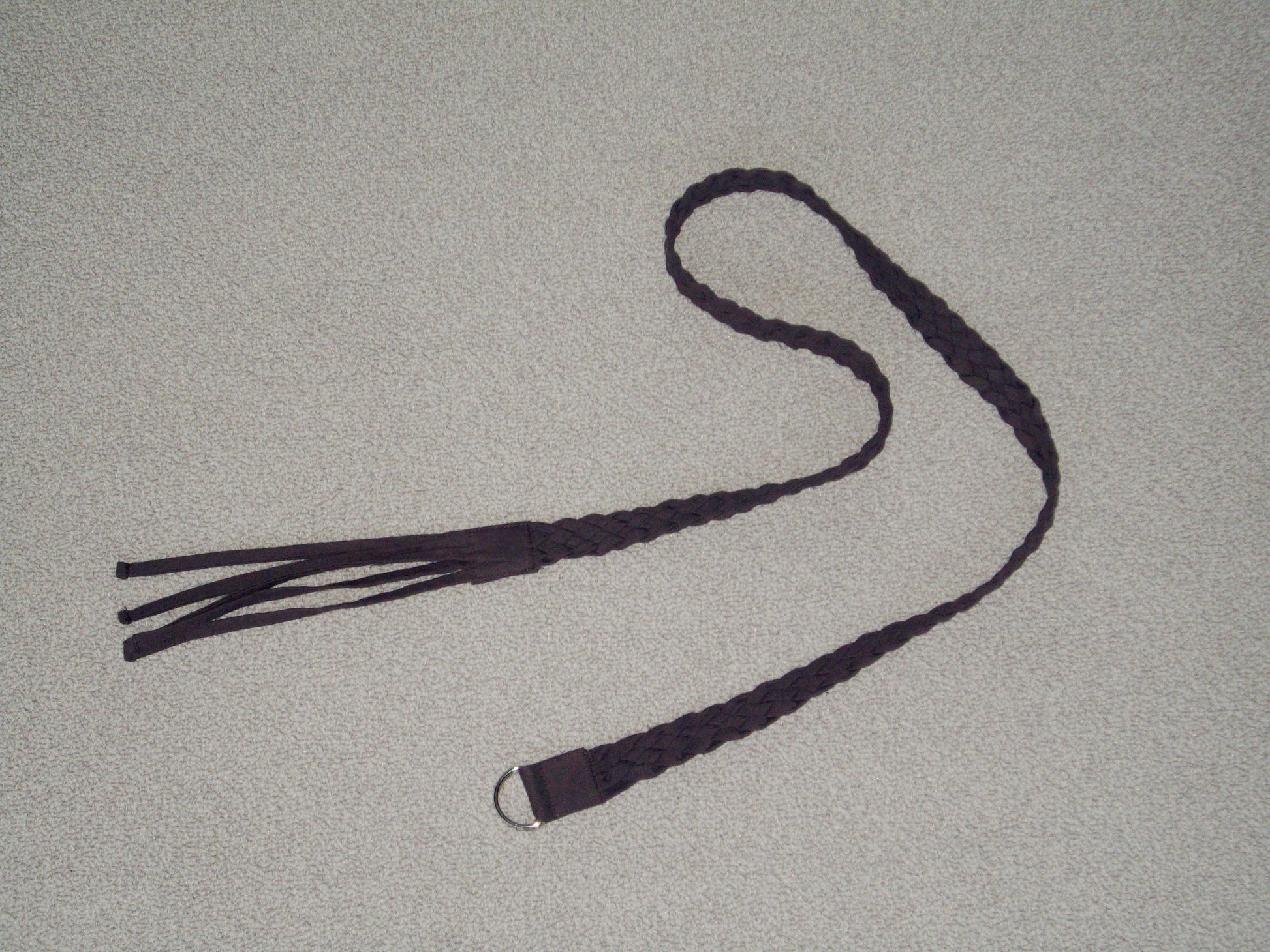 A dark braided leather belt, the Plaited Belt - Chocolate by 0, with fringe on one end and a metal ring on the other, is displayed on a light-colored textured surface.