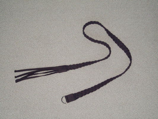 A dark braided leather belt, the Plaited Belt - Chocolate by 0, with fringe on one end and a metal ring on the other, is displayed on a light-colored textured surface.