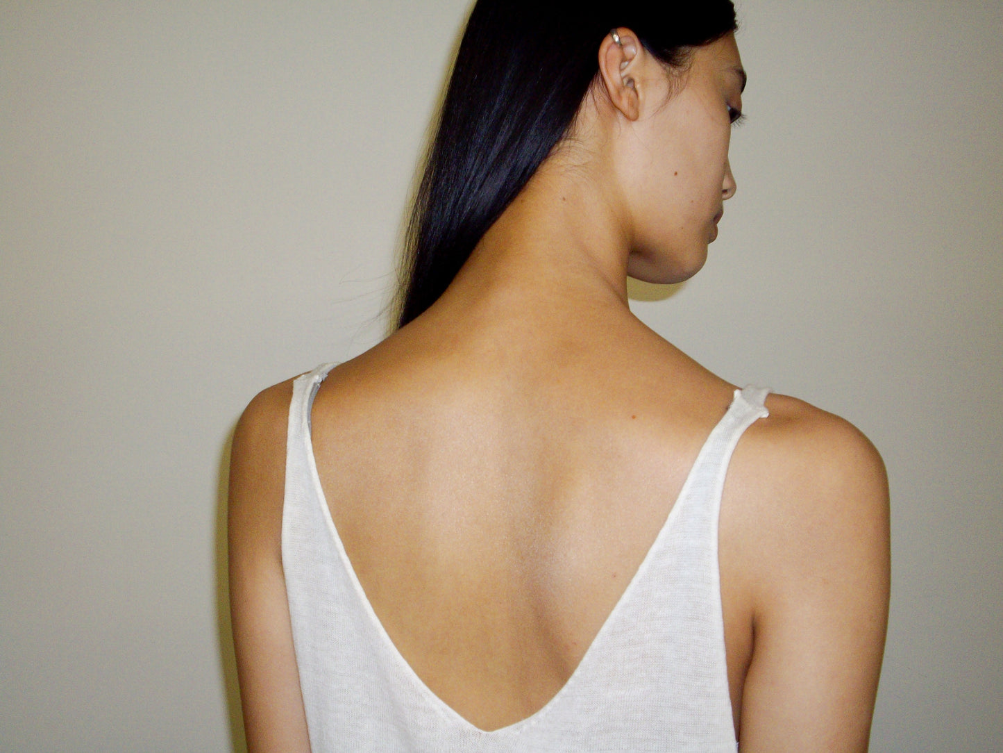 A person with long, straight dark hair is seen from behind, wearing the Rolled Knit Top - Off White by 0, a sleeveless low-back piece, against a plain light background. Their head is slightly turned to the side.