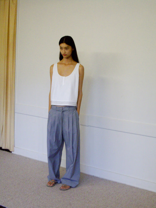 A woman stands indoors in a loose white sleeveless top and Tri Pleat Pure Wool Pant - Steele Stripe with sandals, set against a plain white wall and beige curtain.