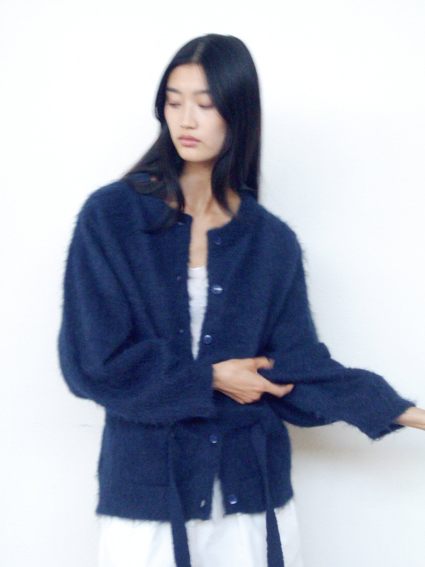 A person with long dark hair models the 0 Belted Cardi - Navy over a white outfit, standing against a plain light background.