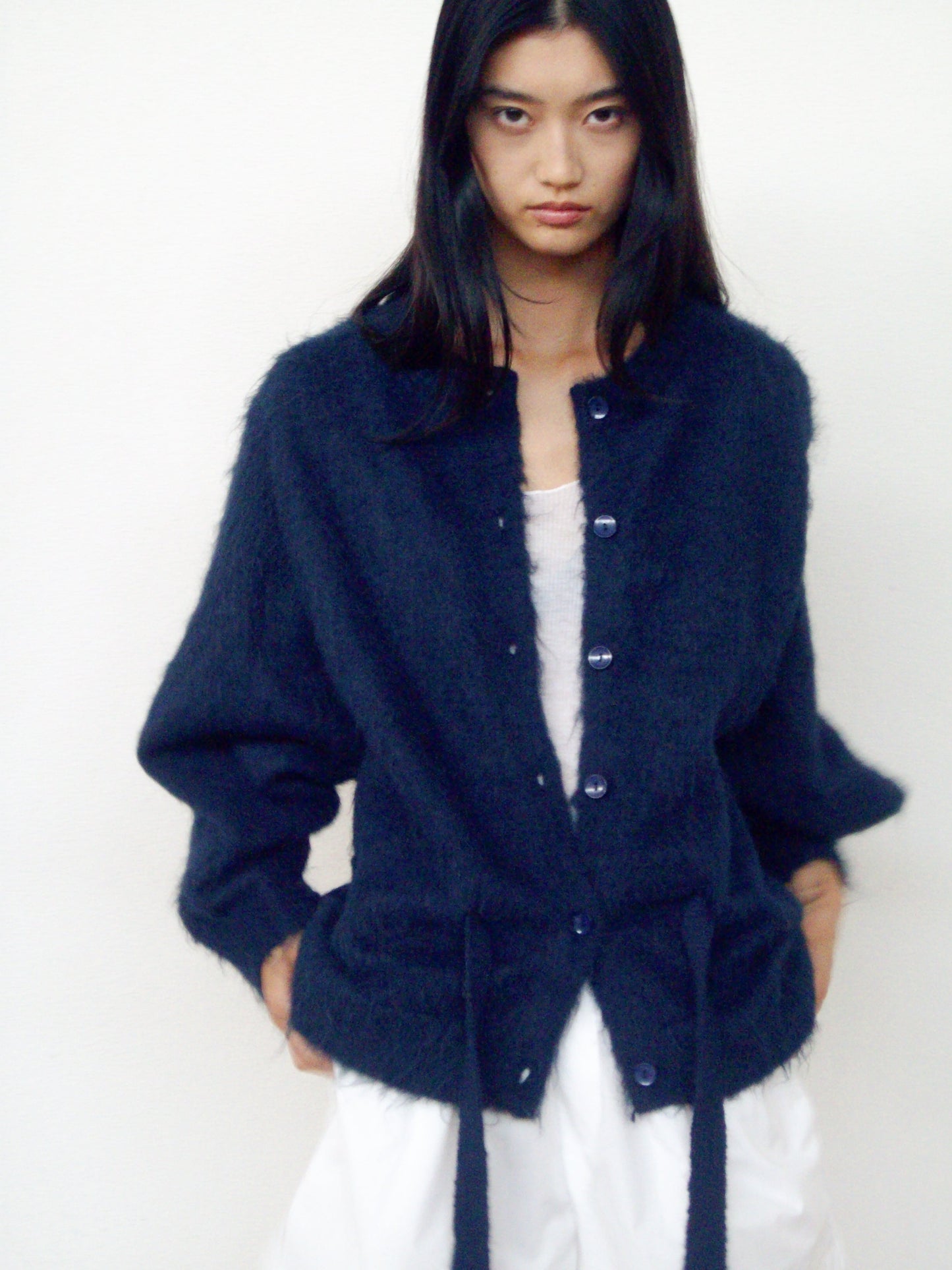 A woman with long dark hair wears the 0 Belted Cardi in Navy over a white top and loose white pants, standing against a plain light background with her hands in her pockets.