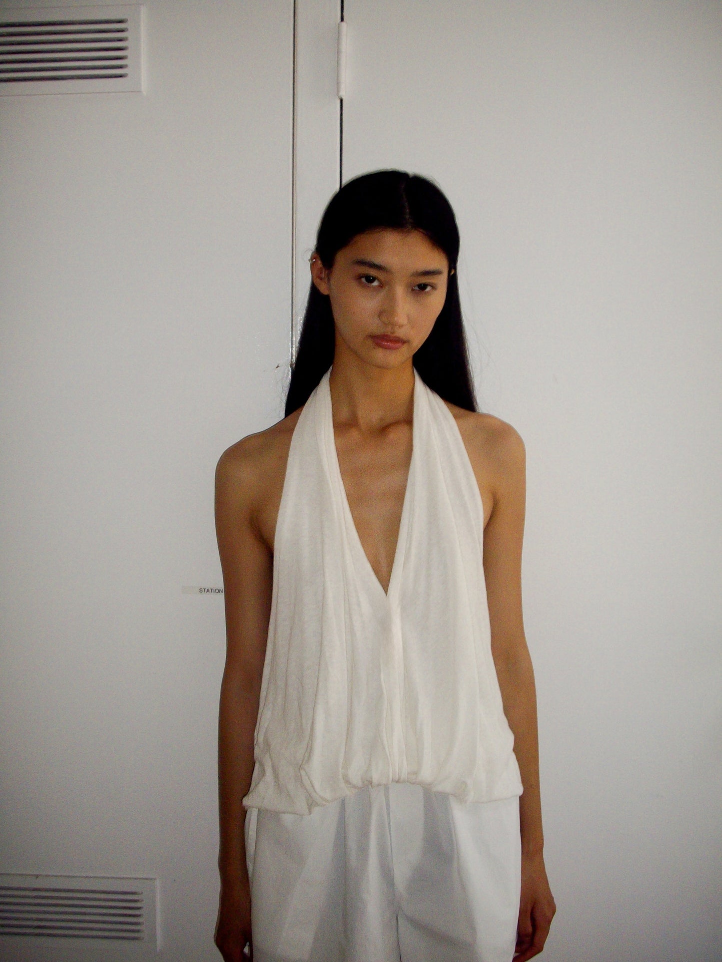 A young woman with long, straight black hair stands against a white background, wearing the 0 Jersey Top in white with a relaxed fit and deep V-neck, paired with loose white pants, looking calmly at the camera.