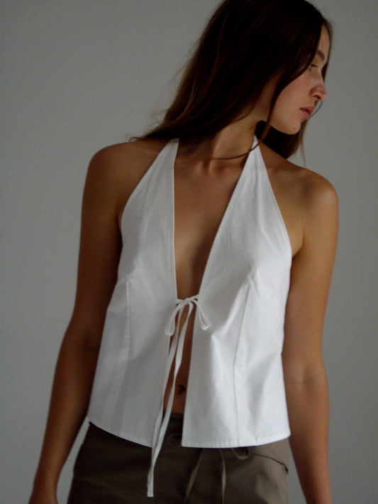 A woman with long brown hair models the Open Halter Top - White, a sleeveless organic cotton halter top with a deep V-neck and front tie, paired with light brown pants against a plain neutral background.