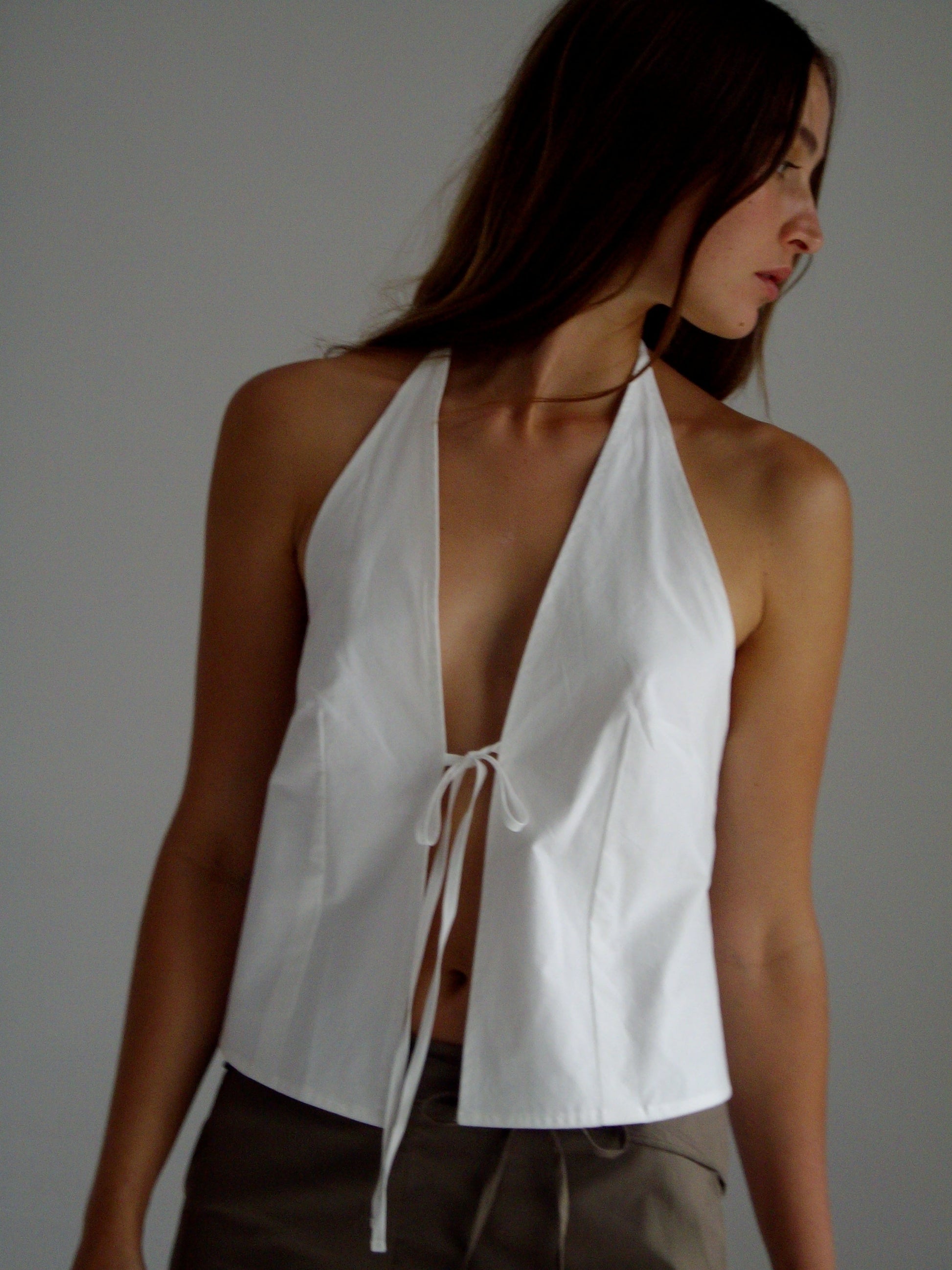 female model wears the open halter top top by Deiji Studios in white

top features open front with centre bust tie, halter neck tie and v neckline, bust seams for subtle shaping and shirred back

made from 100% GOTS certified organic cotton poplin

paired with a cay green cotton trouser