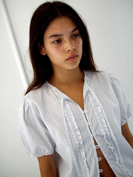 A young woman with long dark hair and freckles wears the Double Frill Top – Sage Stripe from Deiji Studios, standing against a plain white background and looking slightly to the side with a neutral expression.