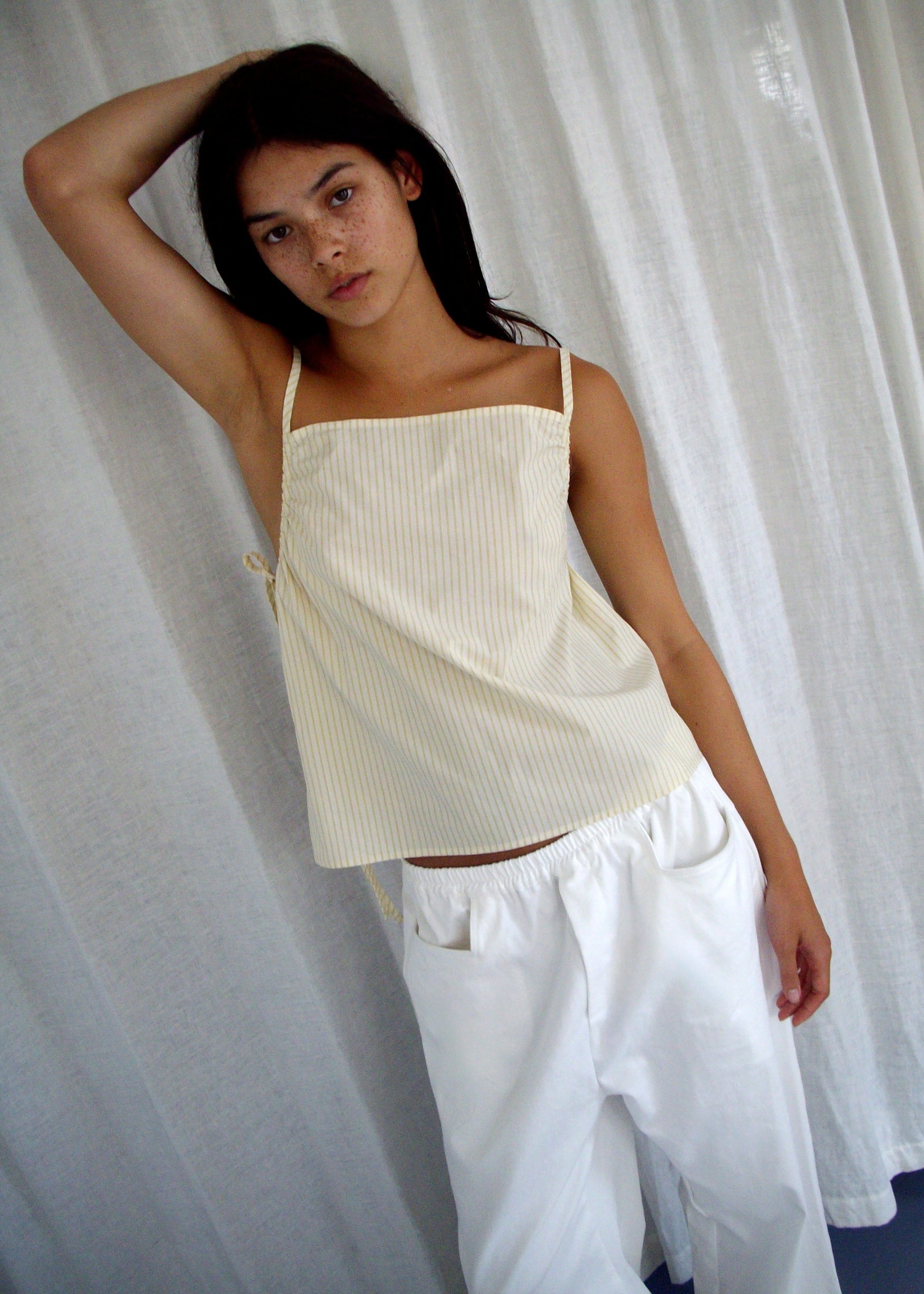 A person with long dark hair poses before white curtains, wearing the Deiji Studios Gathered Strap Top – Butter Stripe in light yellow organic cotton and wide white pants, with their left arm raised and resting on their head.